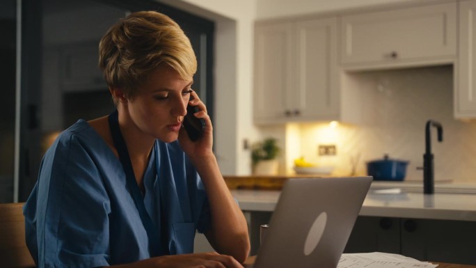 穿着医用工作服的疲惫女人晚上在家里用手机工作或用笔记本电脑学习