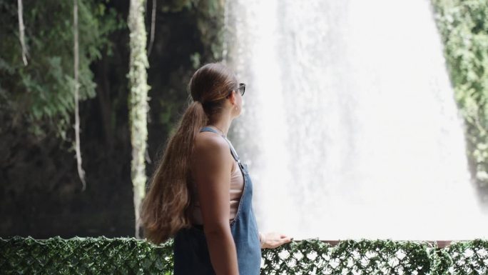 在慢镜头中，一位身穿牛仔连衣裙的年轻女子站在森林中高高的瀑布的背景下环顾四周。