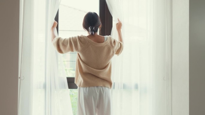 快乐的亚洲女孩与白色的奶油睡衣打开白色的窗帘在窗户上看外面感觉清新与宁静的早晨和新鲜的空气在周末在家