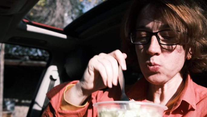 一个45岁的女人坐在驾驶座上，吃着塑料容器里的沙拉