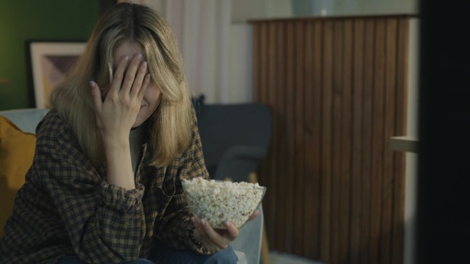 情绪化的年轻女子看恐怖惊悚片表达恐惧，晚上在黑暗的房子里吃爆米花