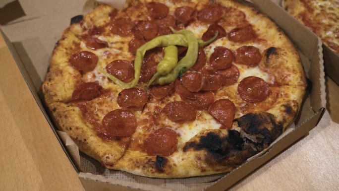 整张意大利辣香肠披萨的俯视图，上面有绿色的辣椒，放在桌子上的纸板披萨盒里