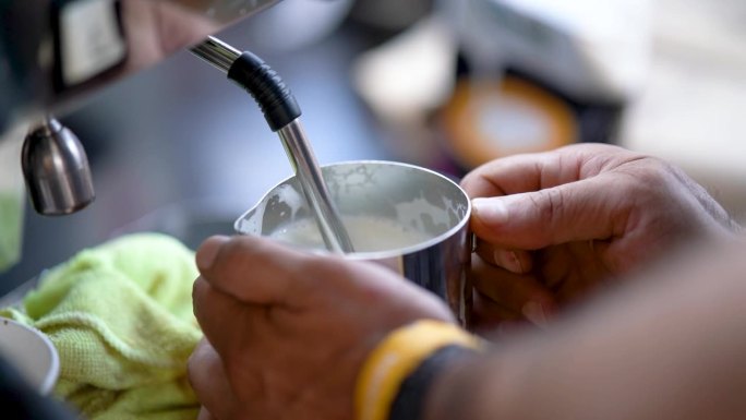 咖啡师在咖啡馆工作的牛奶发泡过程。咖啡师用咖啡机的热蒸汽在大壶里搅打牛奶。制造牛奶泡沫的过程。