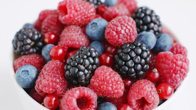夏季概念新鲜覆盆子，黑莓，蓝莓，红醋栗旋转在一个孤立的白色背景顶部视图白色碗。健康的水果浆果。新鲜水
