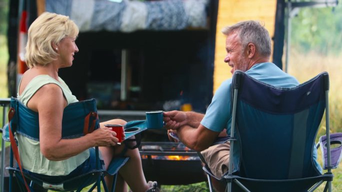 一对老年夫妇开着房车在乡村露营的后景，他们在户外的火炉旁喝咖啡