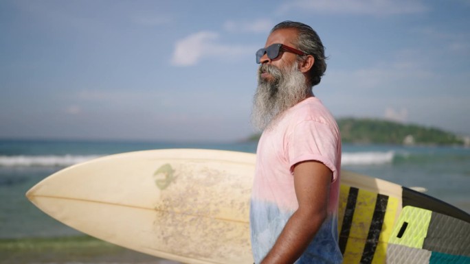 资深旅行者冲浪手拿着冲浪板，在热带岛屿上的异国海滩散步。退休的黑胡子老头戴着墨镜，喜欢旅游，欣赏大海