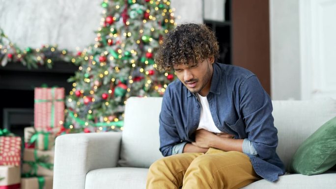 在冬季新年圣诞假期，一个年轻人坐在家里客厅的沙发上感到胃痛
