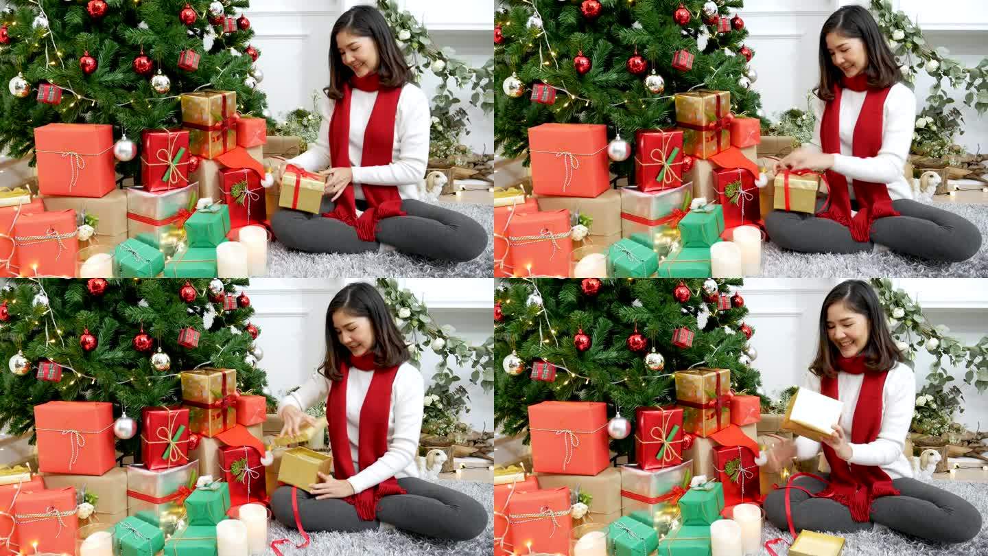 亚洲妇女在圣诞树下打开圣诞礼盒，收到了惊喜礼物。亚洲妇女在客厅里打开礼盒，坐在装饰精美的球圣诞树下庆