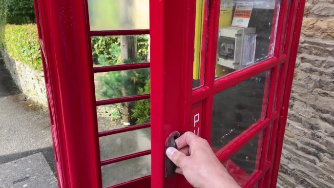 在英国，一个男人的手打开了一个红色电话亭的门，这个电话亭被改造成了一个除颤器亭。