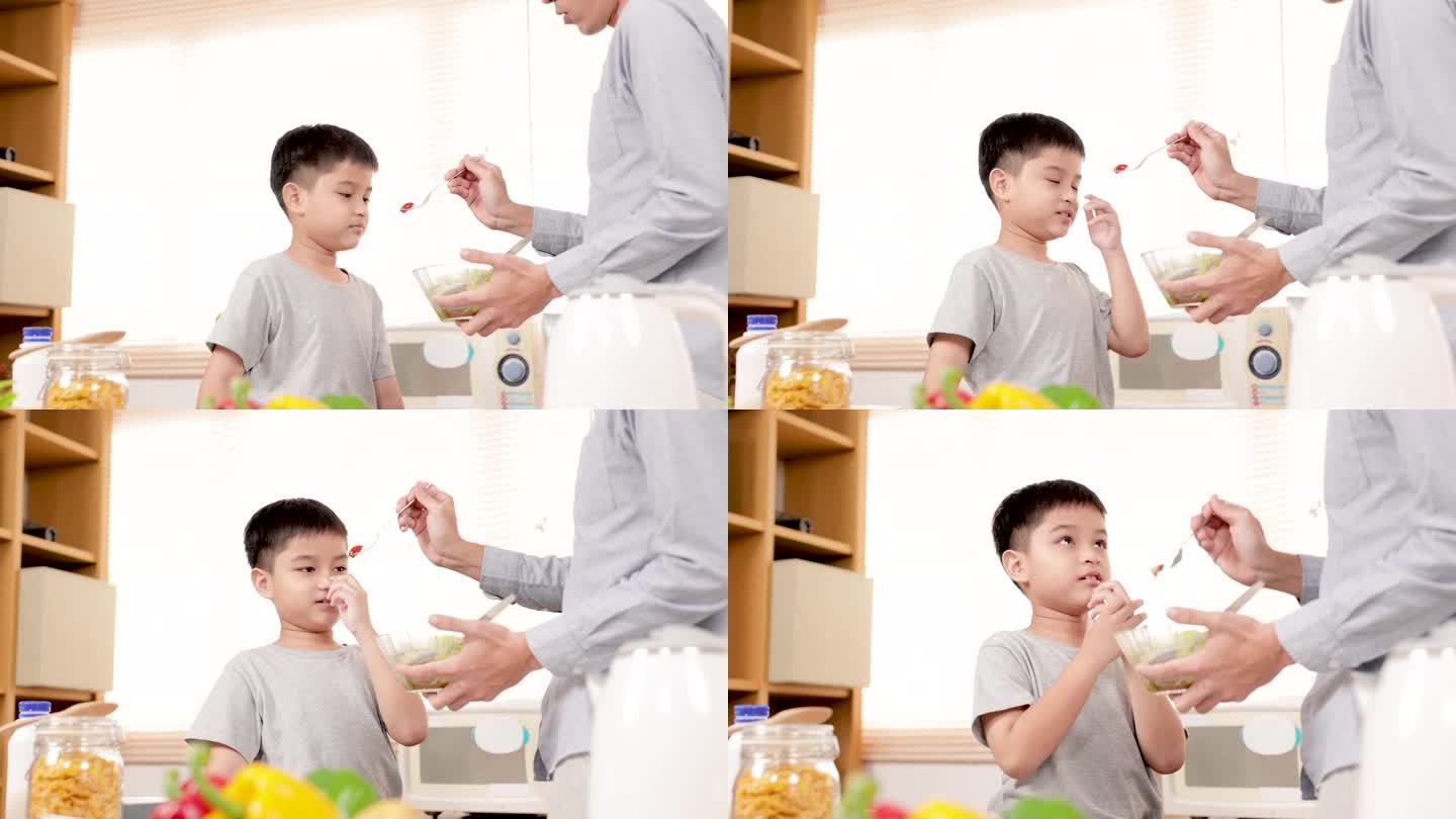家庭活动生活方式在厨房，父亲试图鼓励儿子在厨房吃蔬菜西红柿，不高兴的孩子在碗里恶心的沙拉闭上嘴巴不喜