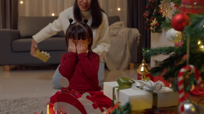 亚洲幸福的家庭享受庆祝新年假期聚会一起在家里晚上。小女孩们坐在地毯地板上，闭着眼睛等着圣诞树旁妈妈送