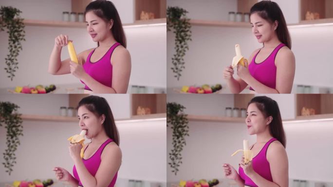 运动的年轻亚洲女性穿着运动服在厨房内吃香蕉，享受吃水果的饮食和健康的运动减肥概念。