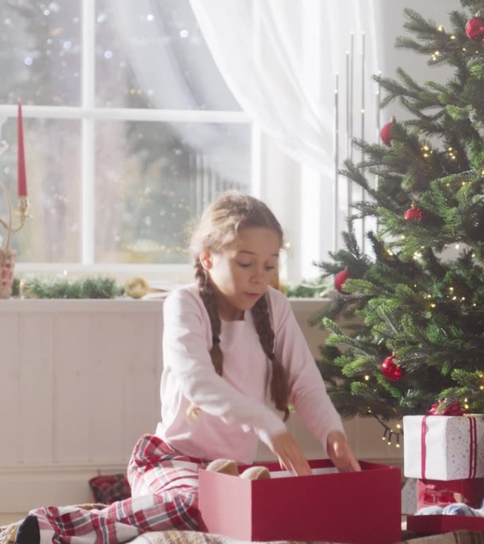 垂直屏幕:快乐的小女孩在节日早晨醒来，从圣诞树下收到新玩具。可爱的孩子迫不及待地从圣诞老人那里拿礼物