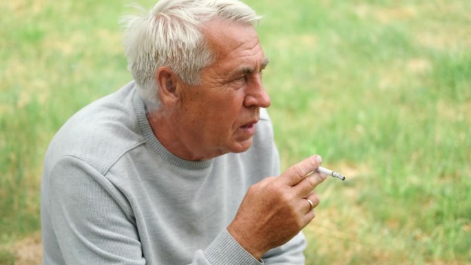 一位老人在户外点燃一支香烟，背景是绿色的大自然。沉思的老人一边抽烟，一边看向一边。特写脸。放松的退休