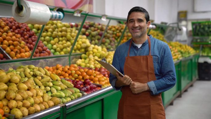 在水果和蔬菜市场工作的成年推销员拿着一个剪贴板，面对着镜头笑得很开心