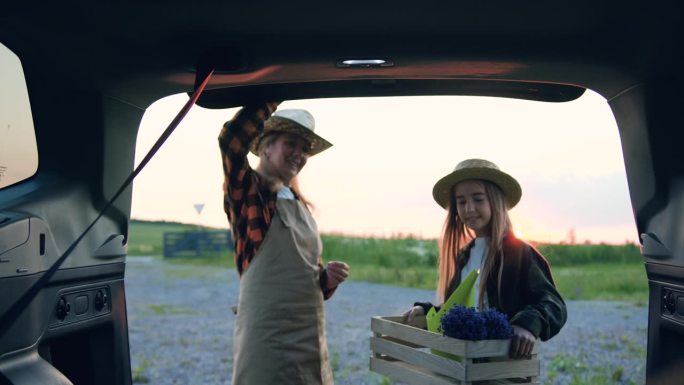 系着围裙的女园丁打开汽车后备箱，她的女儿在夕阳中放了一个装有鲜花的木箱。母亲和戴帽子的女孩从自己的田