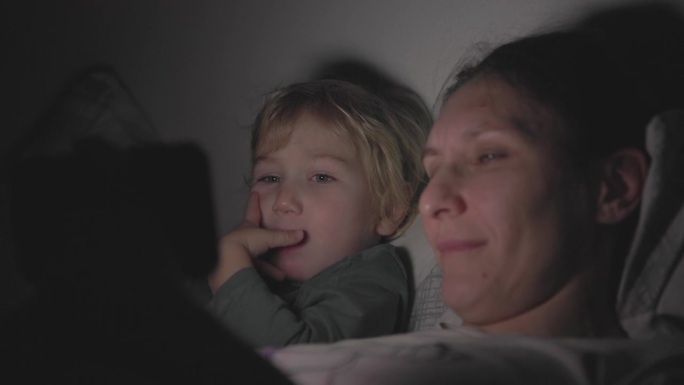 妈妈和儿子关着灯躺在床上，在黑暗的房间里用手机看卡通片