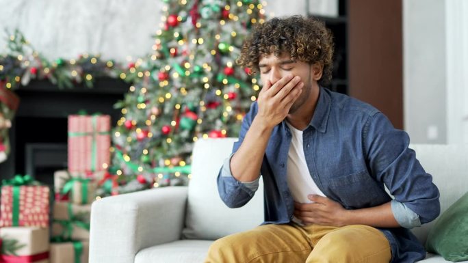 一位年轻英俊的男子在冬季、新年、圣诞假期期间，坐在家里客厅的沙发上恶心得想吐。