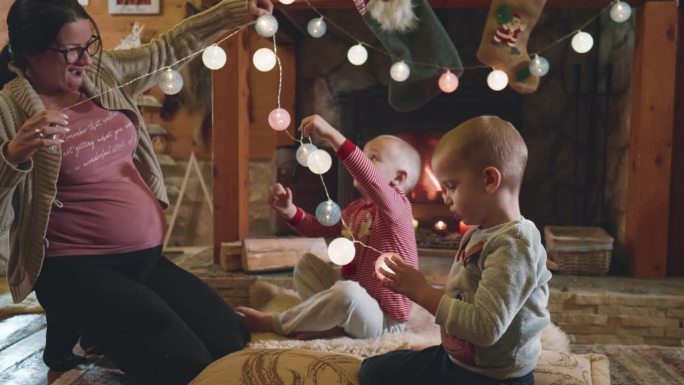 一位怀孕的母亲和两个小孩坐在壁炉旁的地板上，用灯装饰房间，为平安夜做准备
