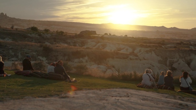 人们坐在山间草地上的靠垫上，看着山后温暖的夕阳。