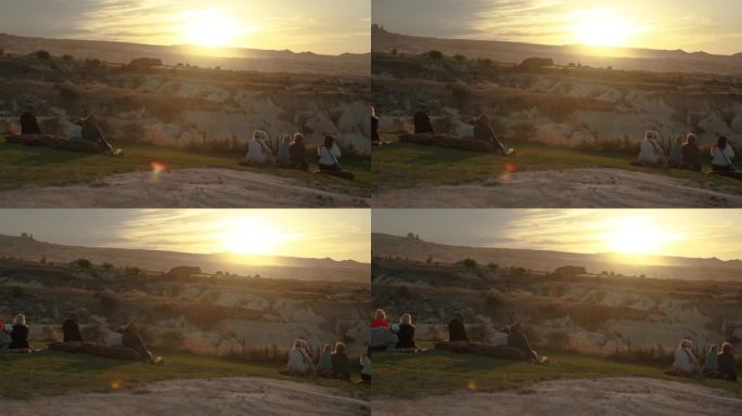 人们坐在山间草地上的靠垫上，看着山后温暖的夕阳。