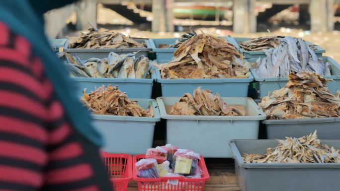 图为白天在印尼传统鱼市出售的鱼、虾、凤尾鱼、鱿鱼干等各种干燥和加工的海产品，背景是停靠在港口的渔船。