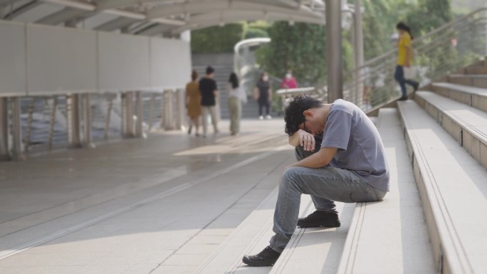 侧视图:一个亚洲人不舒服地坐在车站上，低着头，双手交叉，祈祷奇迹发生在他身上。