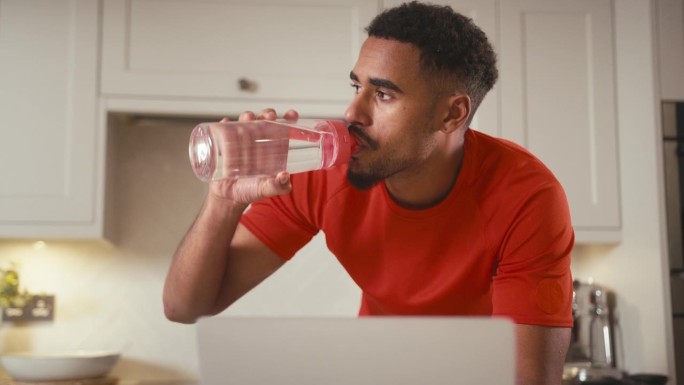 一名男子穿着健身服在家里的厨房里用笔记本电脑喝水