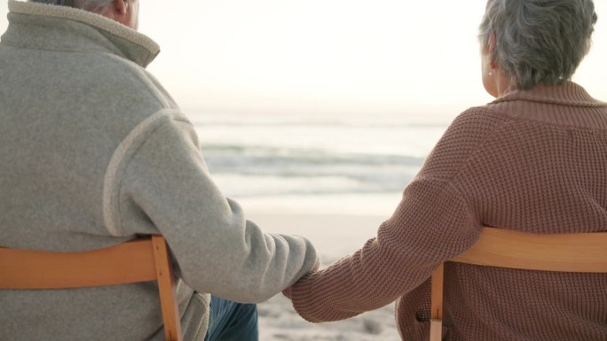 日落、沙滩和老年夫妇坐在海边的椅子上，享受承诺、亲密和美好的时光。爱情，旅行和背影的男人和女人牵着手