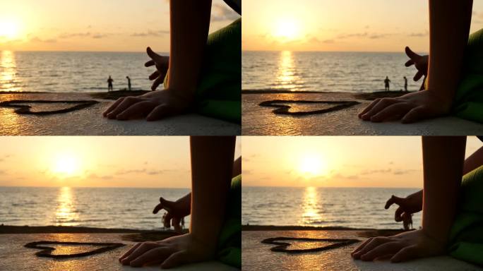 特写，日落时，一个孩子用手指在海滩堤岸人行道的混凝土表面上用沙子画人物。用手指用沙子画爱心