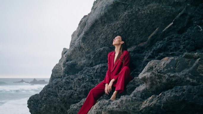 身着优雅红色套装的女子坐在岩壁上。时尚的女孩看起来忧郁的海洋。