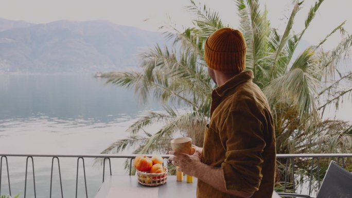 慢镜头:男子端着一杯茶走在阳台上，看着山景湖景
