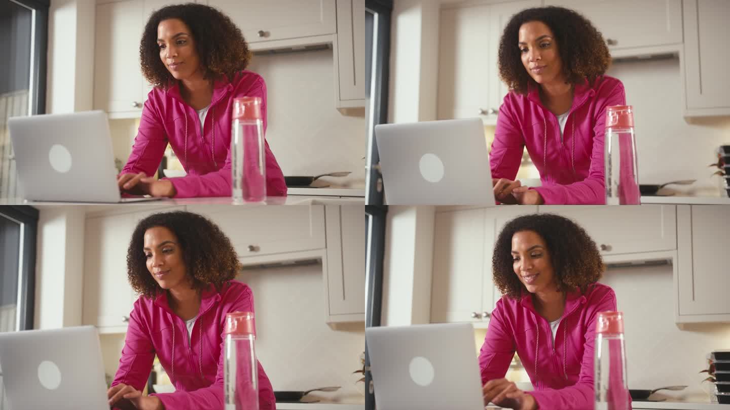 穿着健身服在家里厨房拿着一瓶水在笔记本电脑上工作的女人