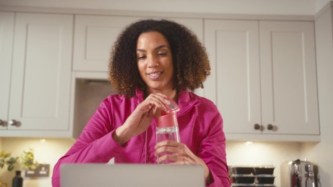 穿着健身服在家里厨房喝瓶装水的女人用笔记本电脑工作