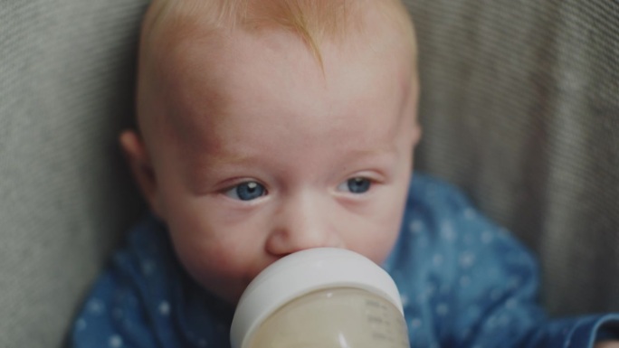 小男孩穿着蓝色的紧身衣坐在椅子上吮吸/喝着一瓶牛奶，小男孩吃东西的特写肖像，婴儿食品概念。饥饿的眼睛
