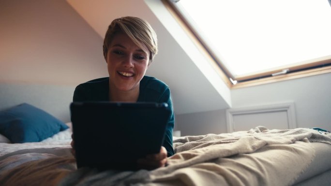 年轻女子穿着睡衣躺在床上对着数码平板电脑大笑