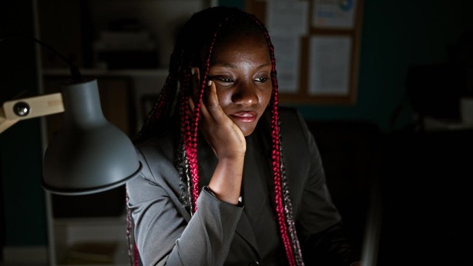 劳累过度的非裔美国女性商务工作者在办公室上晚班，在昏暗的灯光下专注于电脑屏幕。