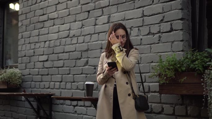 在一条狭窄的街道上，一名年轻女子靠墙站着用智能手机聊天。一个戴眼镜，穿polo衫，手里拿着电话的女孩