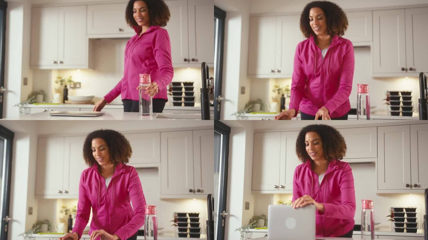 穿着健身服在家里厨房拿着一瓶水在笔记本电脑上工作的女人
