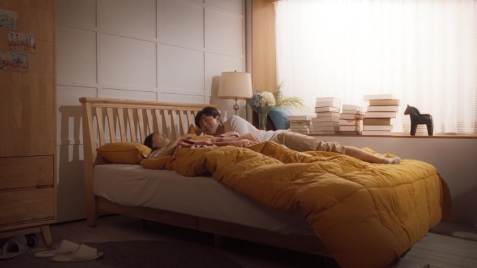 美丽年轻的韩国夫妇睡在床上，阳光照在他们身上，他们看着对方，迎接新的阳光灿烂的一天。两口之家快乐醒来