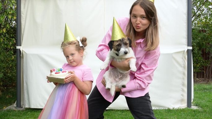 可爱的小女孩和十几岁的女孩在家里的花园里用一个生日蛋糕祝贺她的宠物杰克罗素梗狗。节日庆祝概念。给狗的