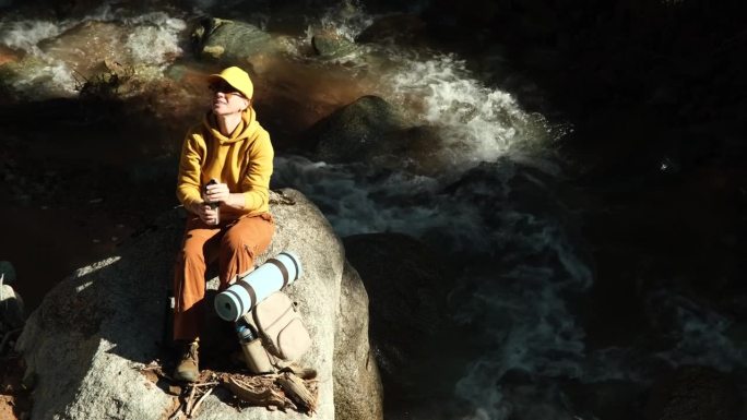 45岁的女人卸下背包，坐在山涧边的石头上，用旅行杯喝水