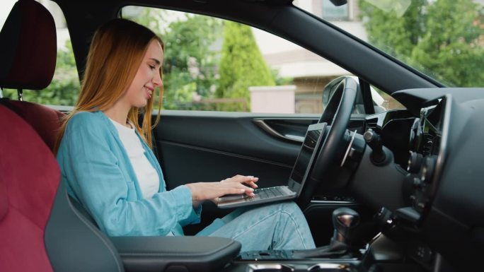 迷人的年轻女子在豪华现代汽车里摆弄笔记本电脑。一位女士坐在驾驶座上，用笔记本电脑上网，收发社交媒体邮