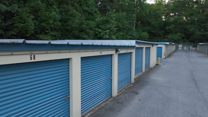 一排带有白色装饰的蓝色储物间，编号的门，一条铺砌的小路，周围环绕着树木。美国自助仓库的空中上升图。