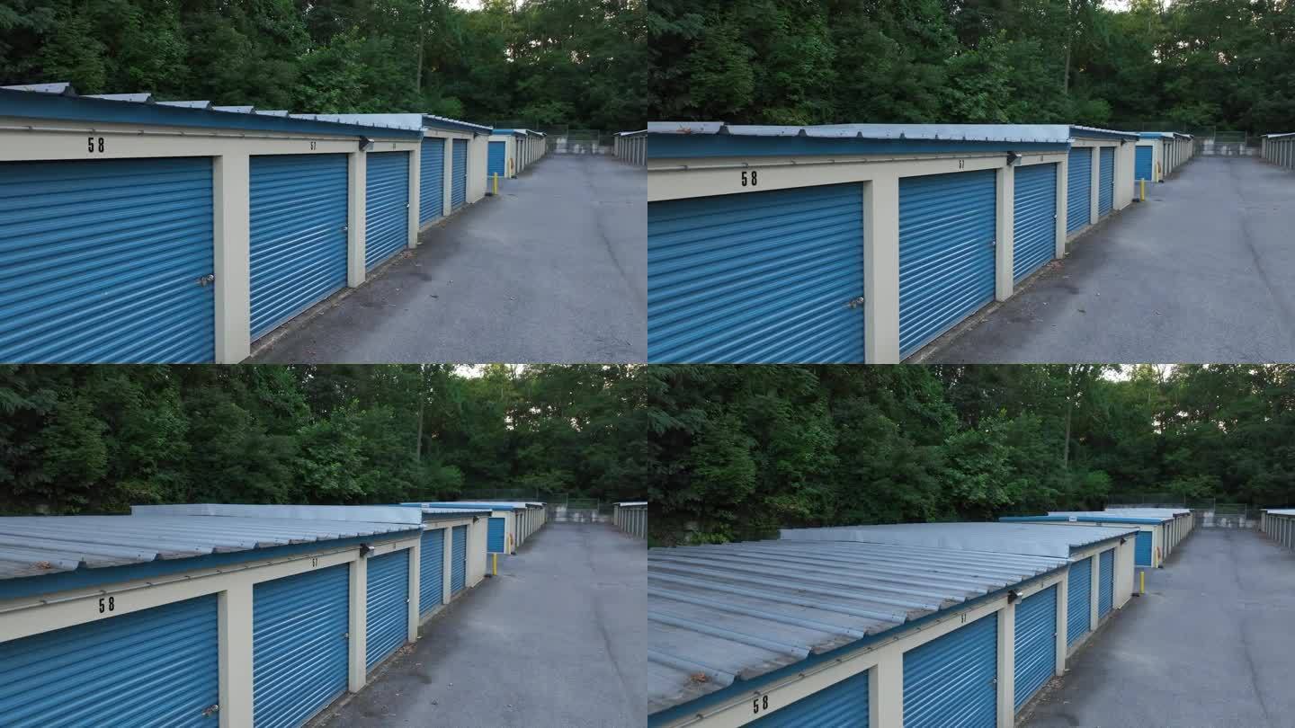 一排带有白色装饰的蓝色储物间，编号的门，一条铺砌的小路，周围环绕着树木。美国自助仓库的空中上升图。