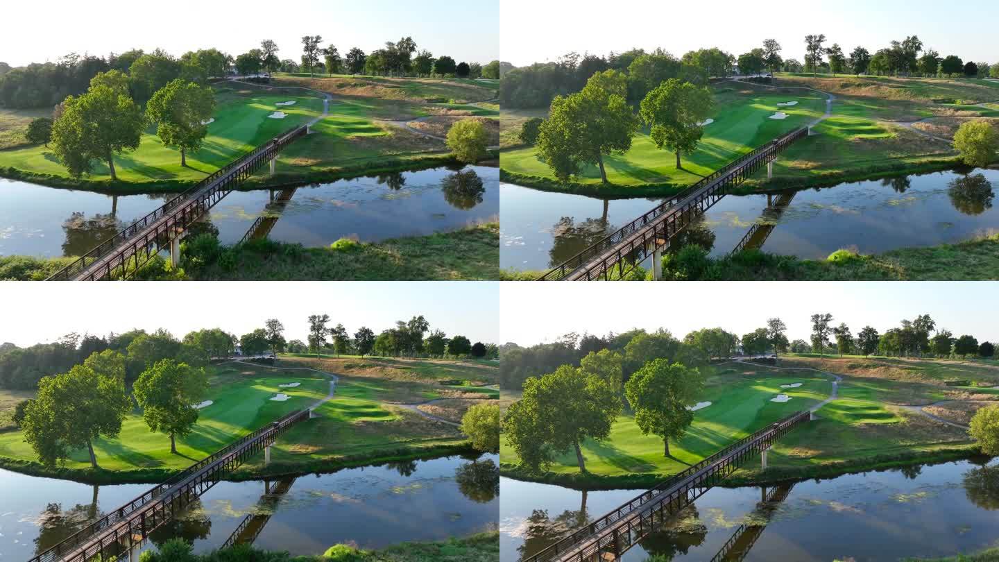 美国阳光明媚的高尔夫球场，以宁静的河流上的木桥、邻近的绿色球道和成熟的树木为特色。日落时高尔夫球车过
