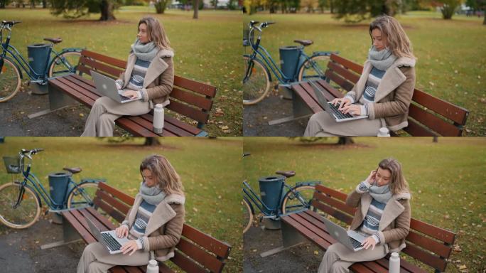 金发女自由职业者穿着时髦的保暖衣服，坐在秋天公园的长椅上使用笔记本电脑