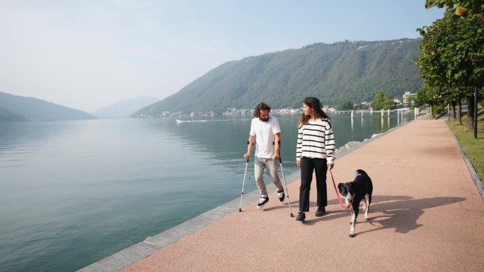一名拄着拐杖的男子与他的伴侣和他们的狗沿着湖上的木板路散步