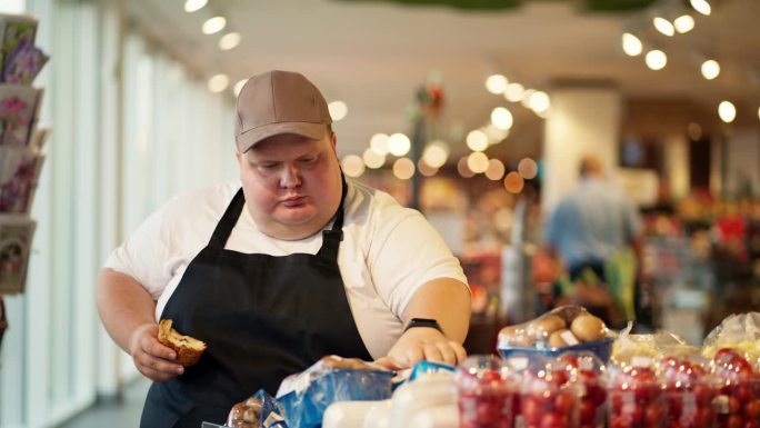 一名穿着白色t恤、系着黑色围裙的超重男性超市工作人员正在吃牛角面包，并在超市柜台上整理食品