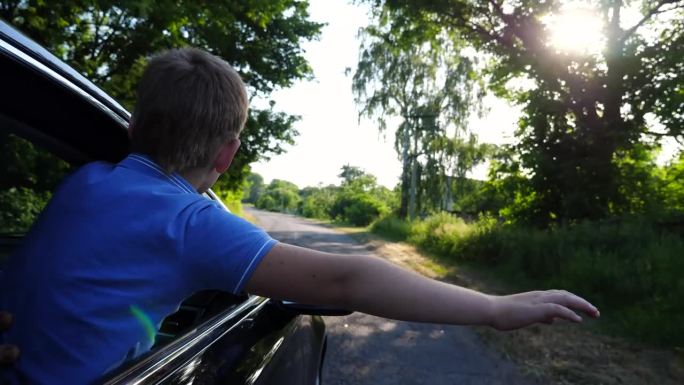 小男孩从汽车车窗探出头来，伸出手，骑着车穿过乡间小路。小孩子在乡下开车享受旅行。旅行的概念。慢动作